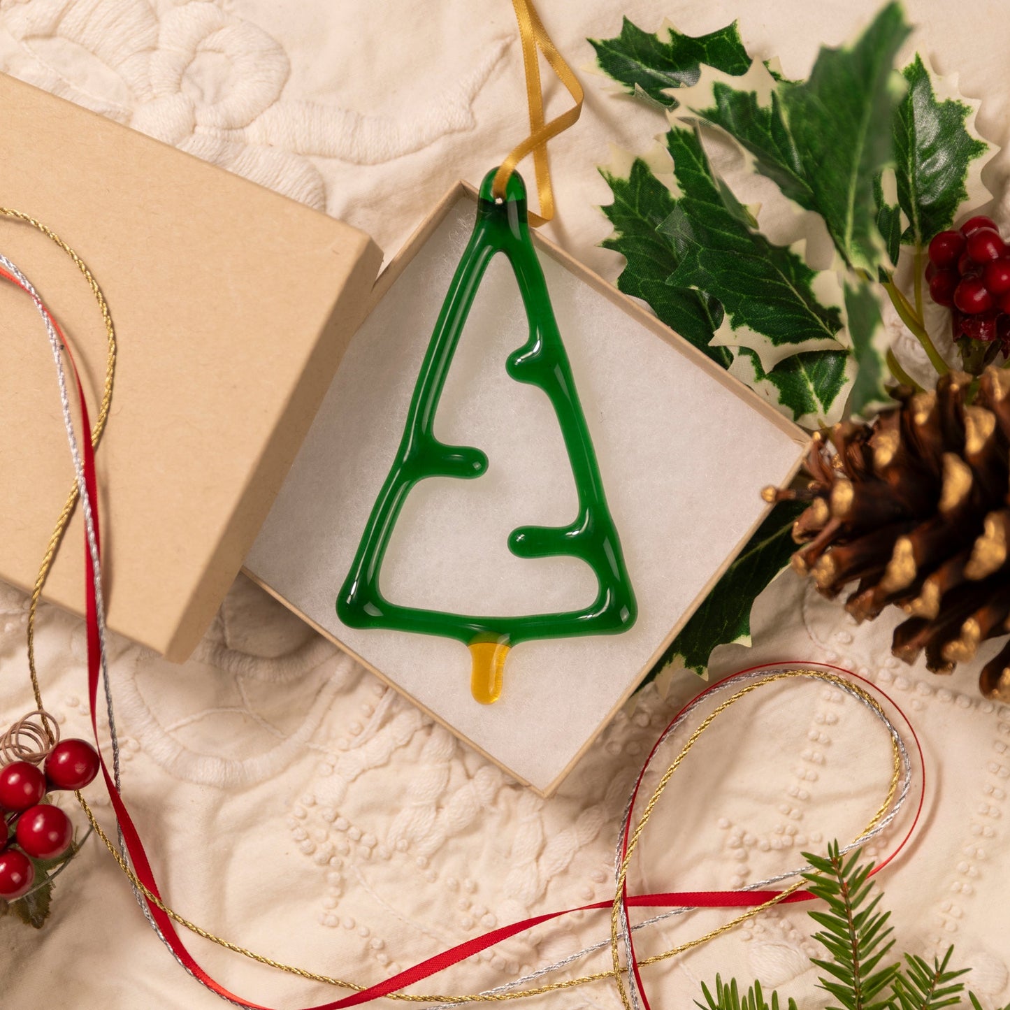 Green Christmas tree ornament in a box with holly leaves, pine cones, and ribbons on a textured surface.