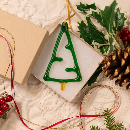 Green Christmas tree ornament in a box with holly leaves, pine cones, and ribbons on a textured surface.